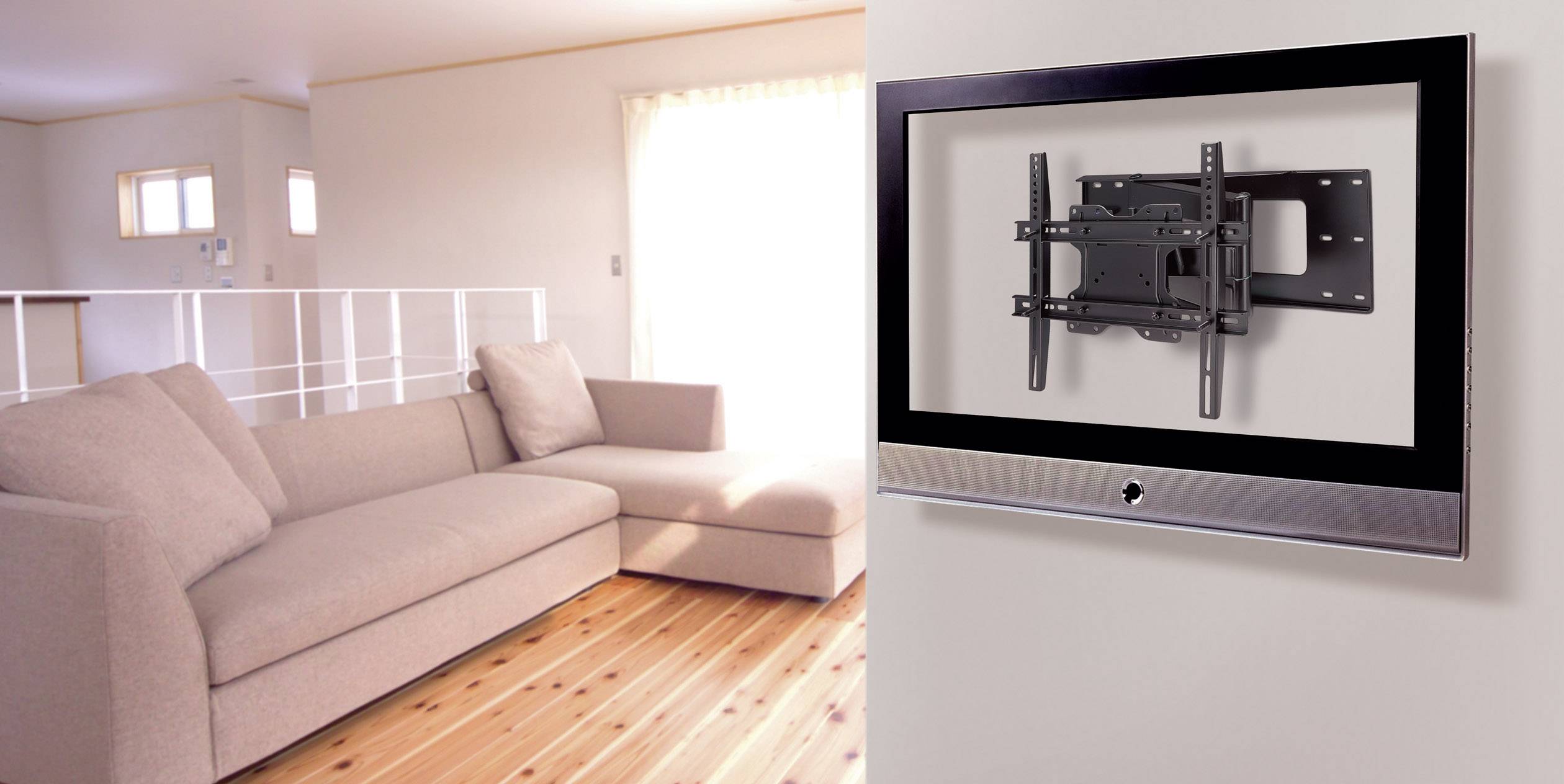 Ein modernes Wohnzimmer mit einem beigen Sofa auf Holzboden. An der Wand eine Fernsehhalterung ohne Fernseher. Helles Tageslicht von einem Fenster.