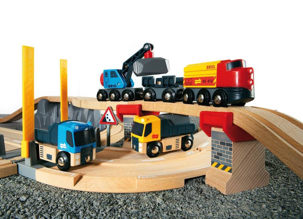 Spielzeug-BRIO-Eisenbahnset mit einem rot-gelben Güterwagen, blauem Kranwagen und zwei kleinen Lastwagen auf Holzgleisen und einer Brücke.