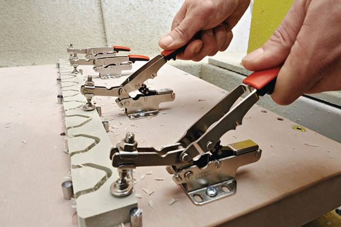 Hände, die Spannzangen verwenden, um Metallbeschläge auf einer Platte zu befestigen, mit mehreren Spannzangen in einer Reihe auf einer Werkbank.