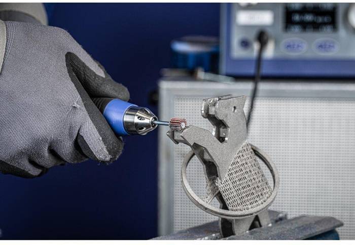 Eine Person trägt einen grauen Handschuh und verwendet ein Dremel-Werkzeug an einem Metallstück, was einen Prozess des Verfeinerns oder Formgebens des Materials andeutet.