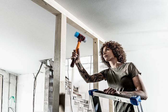 Eine Person auf einer Stehleiter, die mit einem Hammer an einem Holzrahmen während einer Hausrenovierung in einem teilweise fertiggestellten Raum arbeitet.