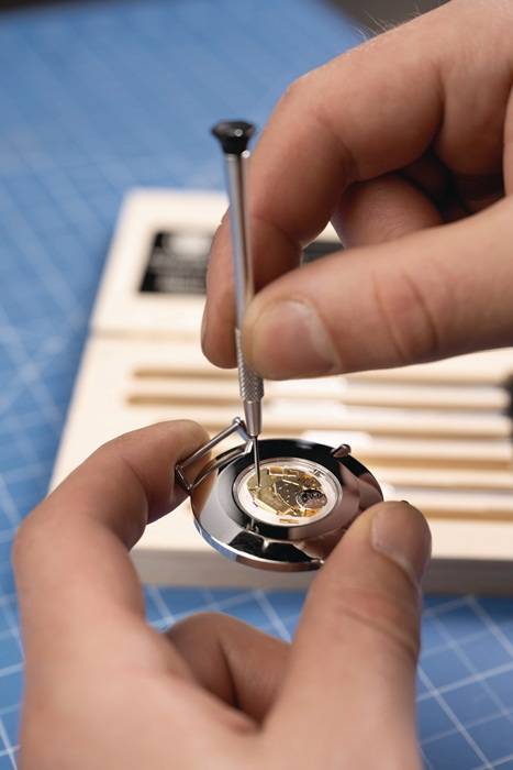 Eine Person, die einen Uhrmechanismus mit einem kleinen Werkzeug sorgfältig zusammenbaut und sich auf die komplexen Zahnräder konzentriert, auf einer blauen Gitternetzarbeitsfläche.