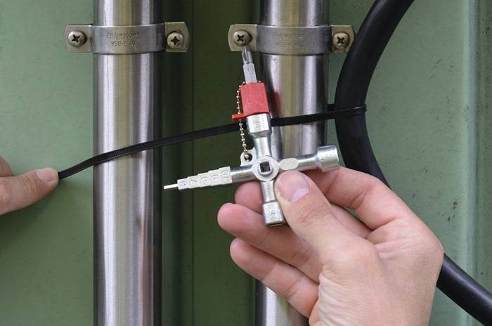 Eine Hand hält einen Vierwege-Multifunktionsschlüssel in der Nähe von zwei gesicherten Rohren, höchstwahrscheinlich für Wartungs- oder Einstellarbeiten in einem industriellen Umfeld.