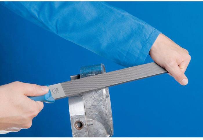 Eine Person mit einem blauen Ärmel feilt ein Metallobjekt mit einer großen, flachen Feile vor einem blauen Hintergrund.