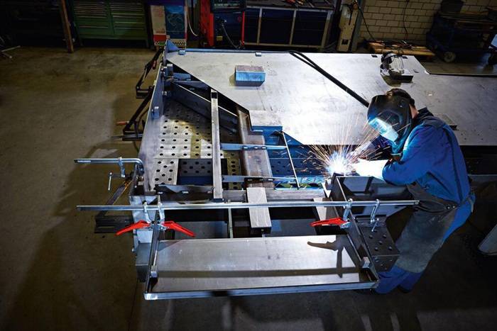 Ein Schweißer in blauer Schutzausrüstung schweißt einen großen Metallrahmen auf einer Werkbank, mit hellen Funken und Werkstattausrüstung im Hintergrund.