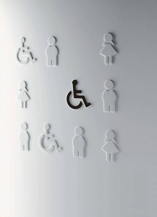 Symbole, die in einem Raster an einer Wand angeordnet sind und zugänglichkeits- sowie Geschlechtersymbole zeigen. Ein Symbol, das Rollstuhlsymbol, sticht durch seine dunklere Färbung besonders hervor.