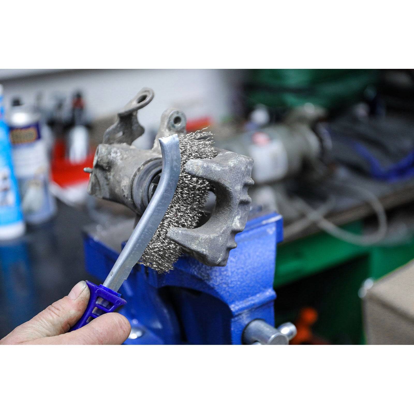 Eine Person reinigt ein Fahrradkettenblatt mit einer Drahtbürste in einer Werkstatt, eingespannt in einem blauen Schraubstock, mit Werkzeugen im Hintergrund.