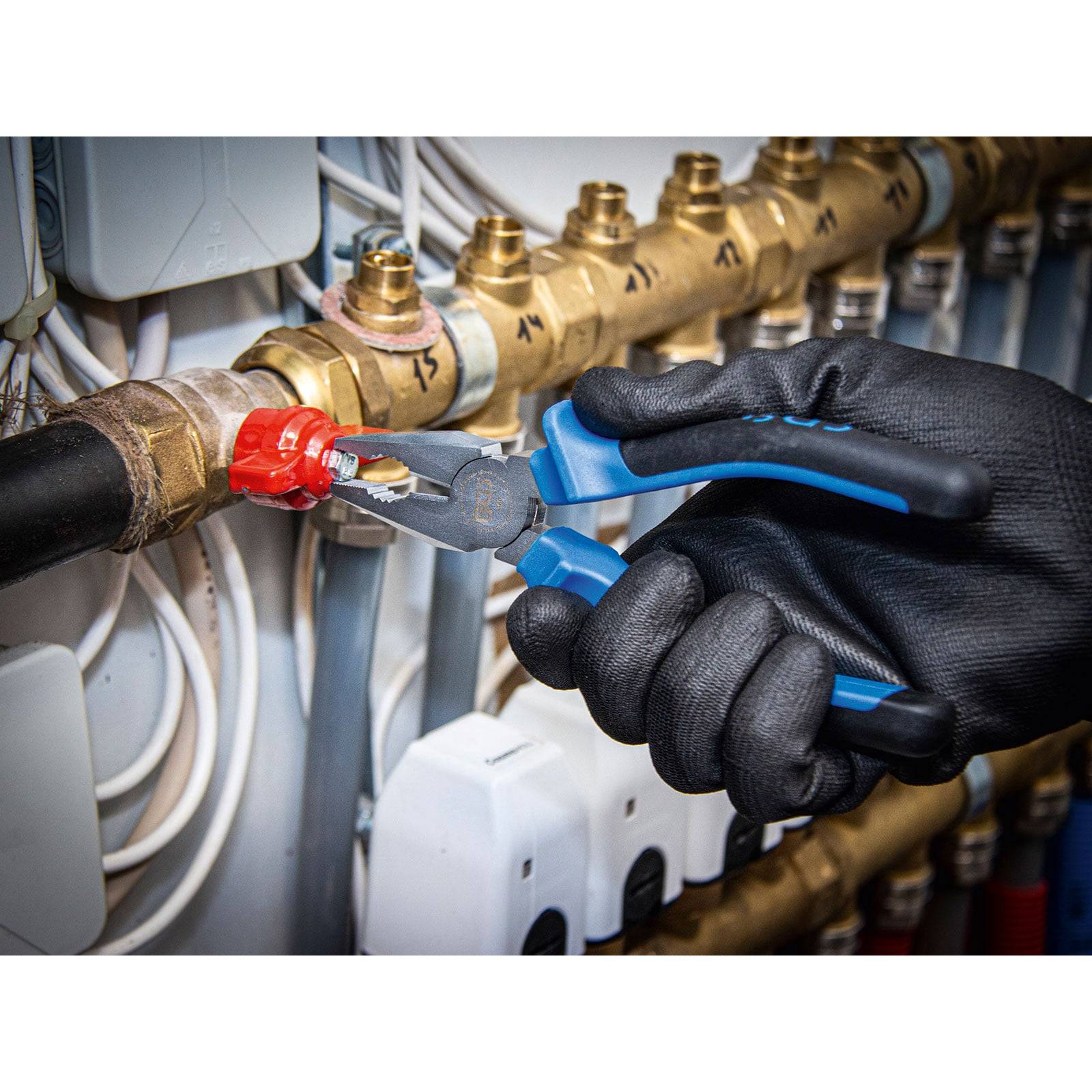 Eine Hand mit Handschuh benutzt eine Zange, um ein rotes Ventil in einem Rohrsystem einzustellen, umgeben von mehreren Rohren und Ventilen vor einer Kontrollleiste.