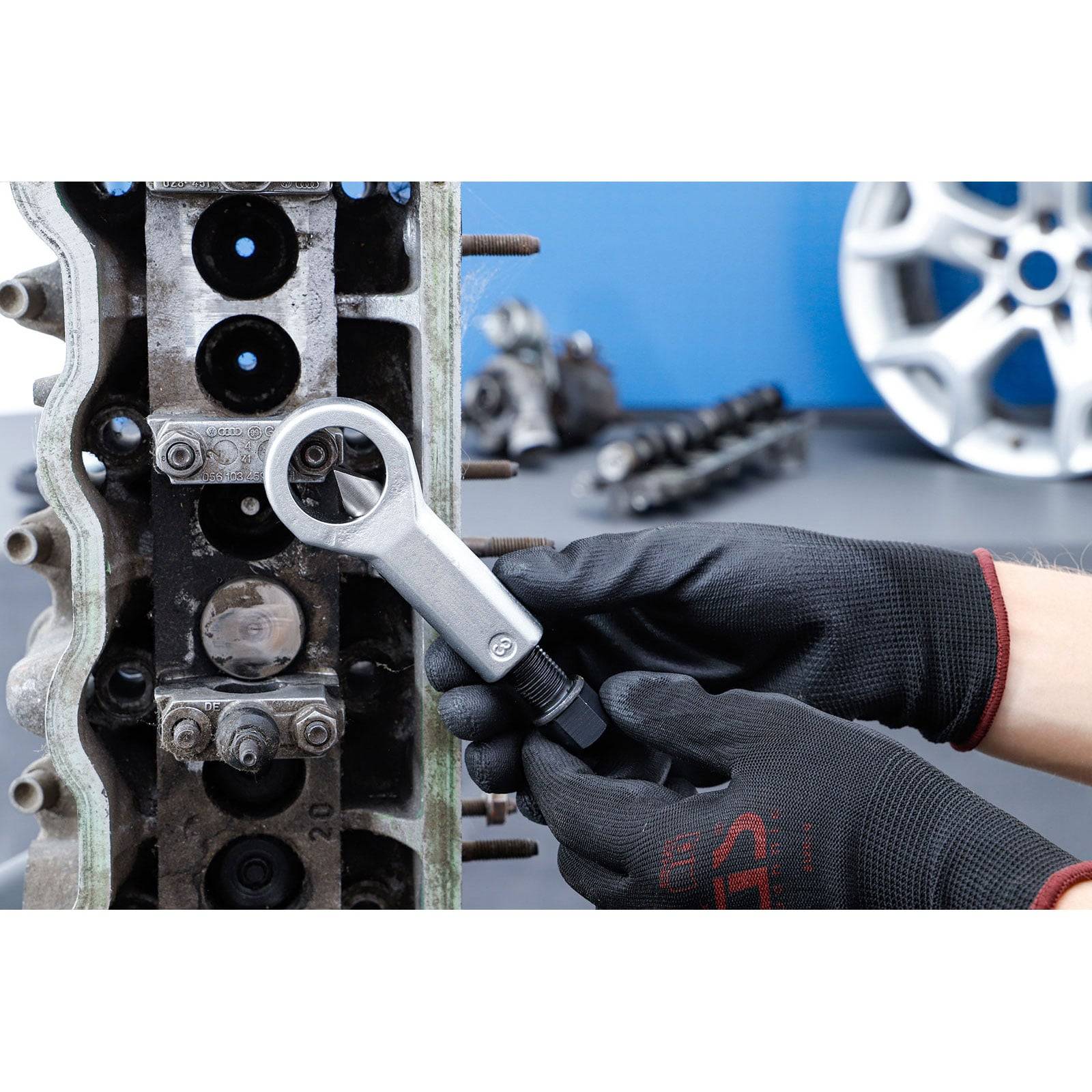 Mechaniker mit schwarzen Handschuhen, der einen Drehmomentschlüssel an einem Motorblock verwendet, wobei Automobilteile im Hintergrund sichtbar sind und Reparaturarbeiten zeigen.