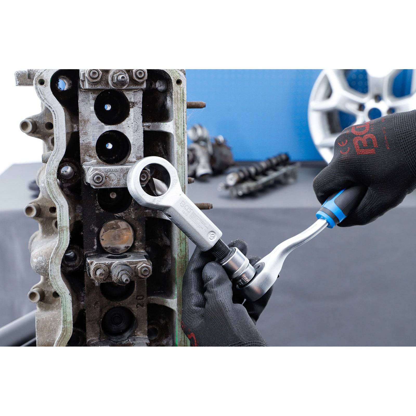 Ein Mechaniker benutzt einen Schraubenschlüssel, um eine Schraube am Motorblock zu befestigen und trägt dabei Handschuhe. Im Hintergrund sind Autoersatzteile und ein Reifen zu sehen.