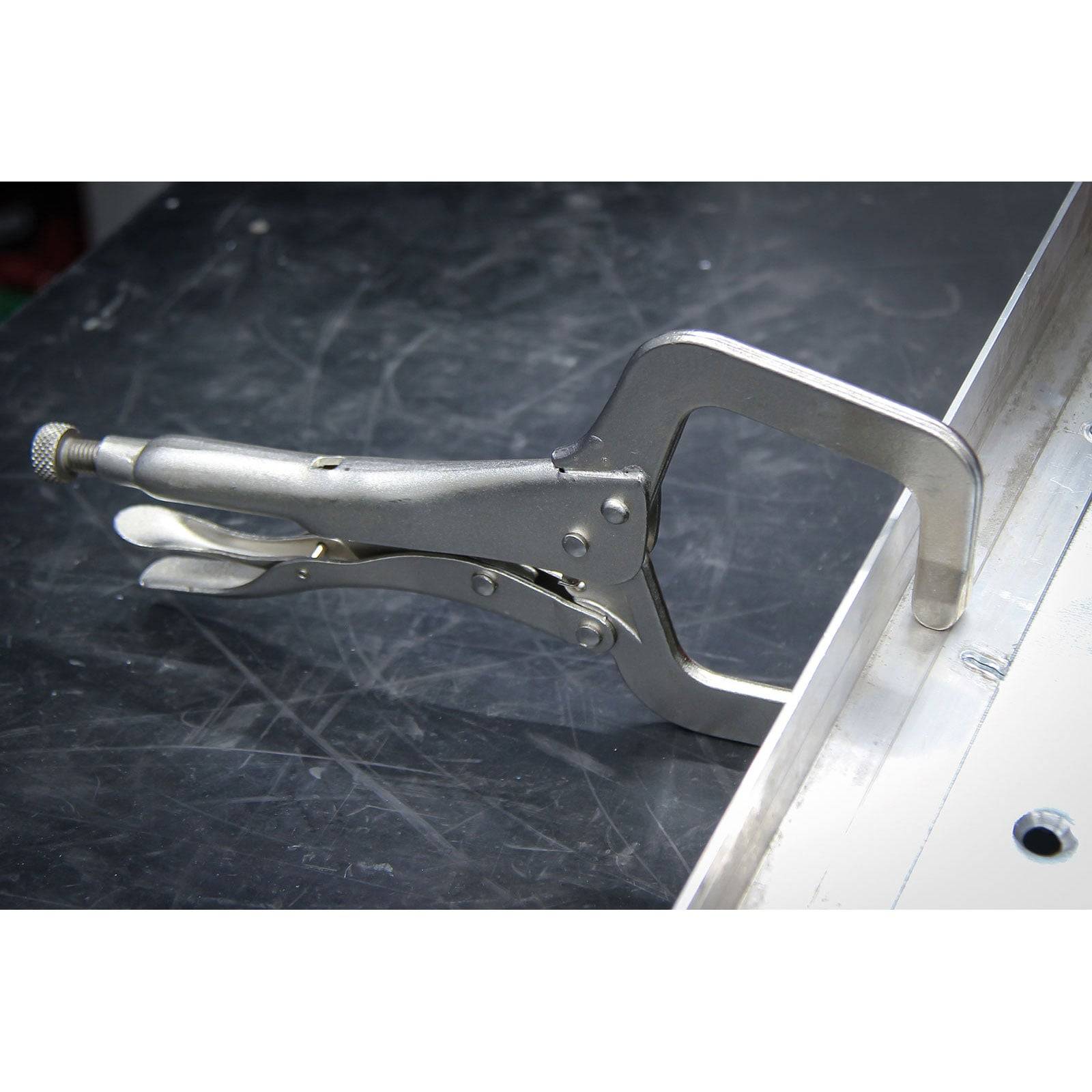 Eine Metallschraubzwinge mit C-Profil ist an einem flachen Metallstück auf einer dunklen, glatten Oberfläche befestigt und demonstriert deren Klemmfunktion.