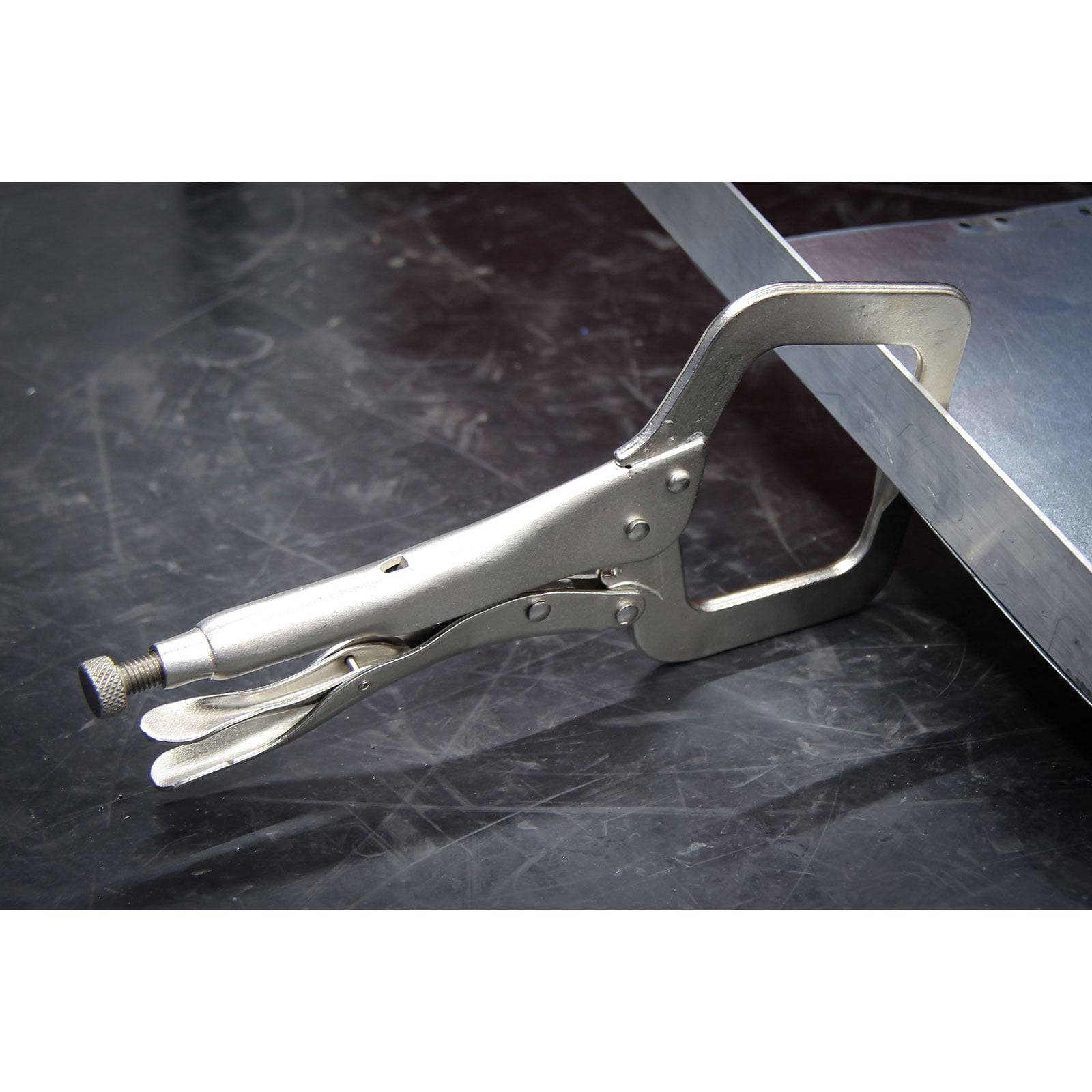 Eine metallische C-Klemme umfasst fest den Rand eines Metallblechs auf einer dunklen Werkbankoberfläche und zeigt damit ihre Verwendung beim Befestigen von Materialien.