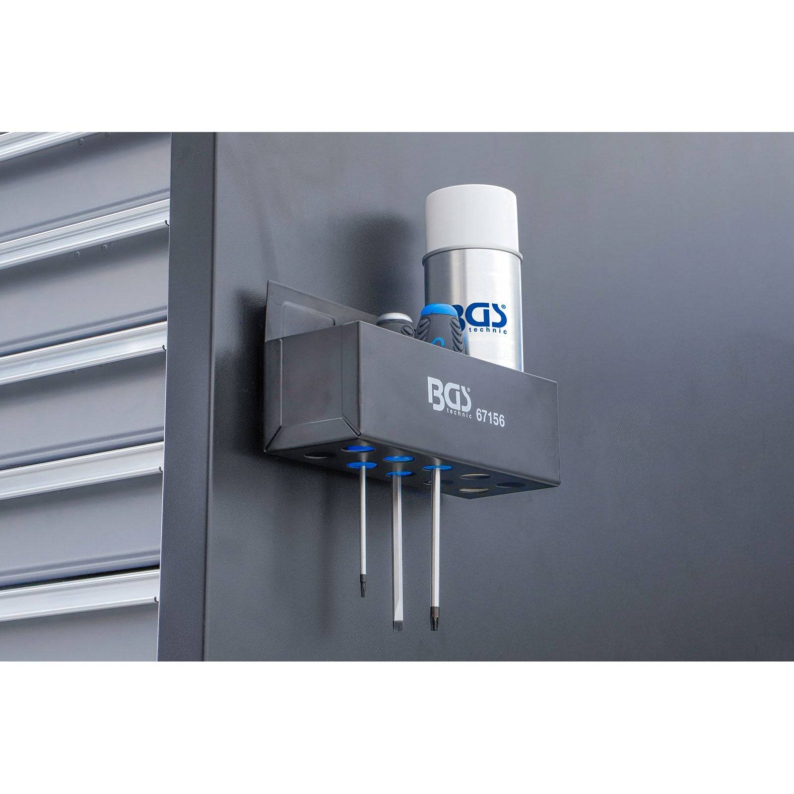 Ein an der Wand befestigter magnetischer Werkzeughalter mit zwei Schraubenziehern und einer Sprühdose, neben metallischen Schubladen, in einer Werkstattumgebung.