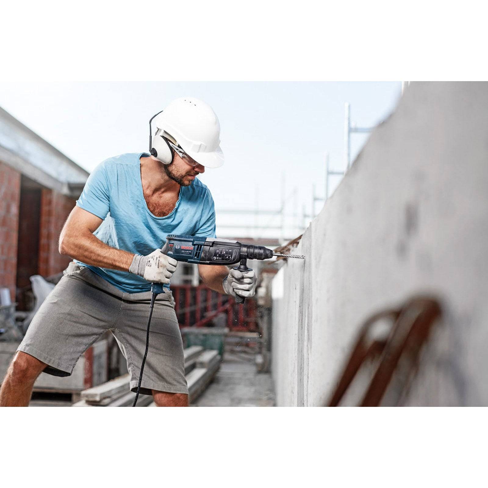Ein Bauarbeiter in einem blauen Hemd und Helm benutzt einen Akkuschrauber an einer Betonwand auf einer Baustelle. Werkzeuge und Materialien sind zu sehen.