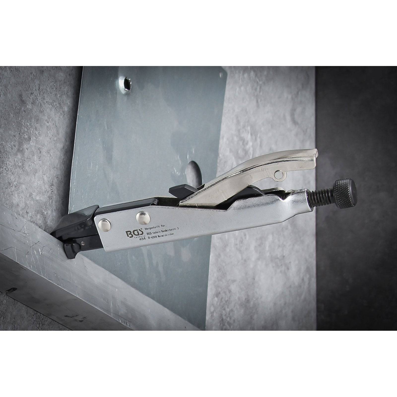 Eine Metallklemme hält ein flaches Metallstück gegen eine strukturierte Wand zur Unterstützung oder Montage. Die Klemme ist teilweise angezogen.