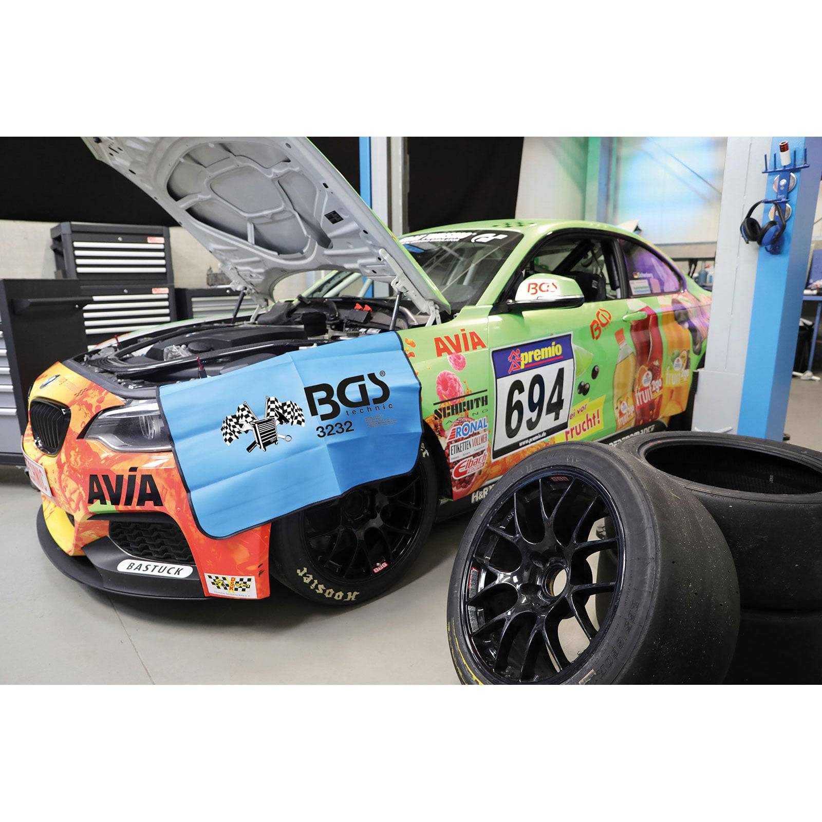 Ein Rennwagen mit der Nummer 694, die Motorhaube geöffnet, steht in einer Garage. Er ist farbenfroh mit verschiedenen Logos und Farben geschmückt. In der Nähe befinden sich Ersatzreifen.