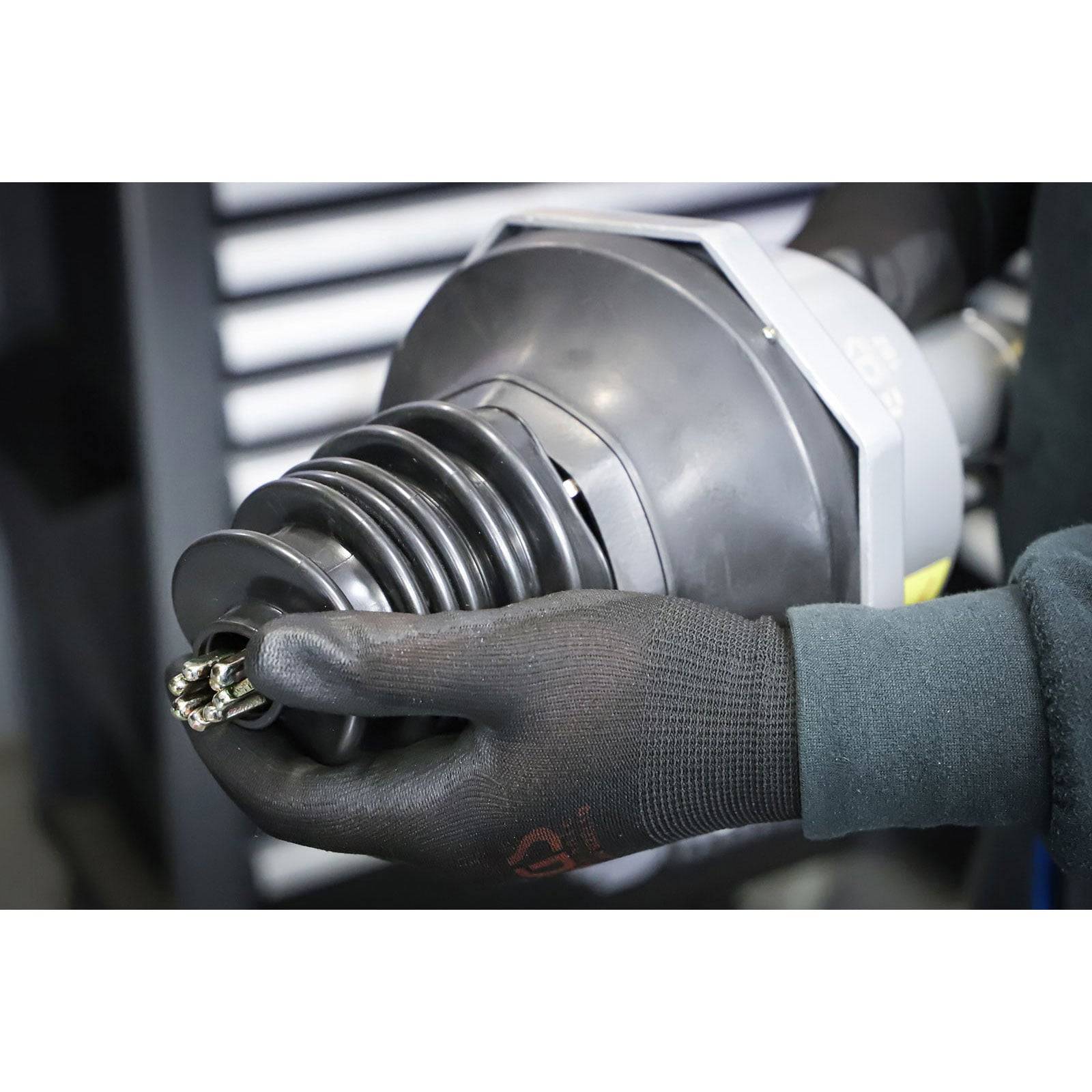 Eine Arbeiterhand mit Handschuh, die eine große mechanische Komponente mit schwarzen Gummirillen hält, die offenbar Teil einer industriellen oder automobilen Montage ist.