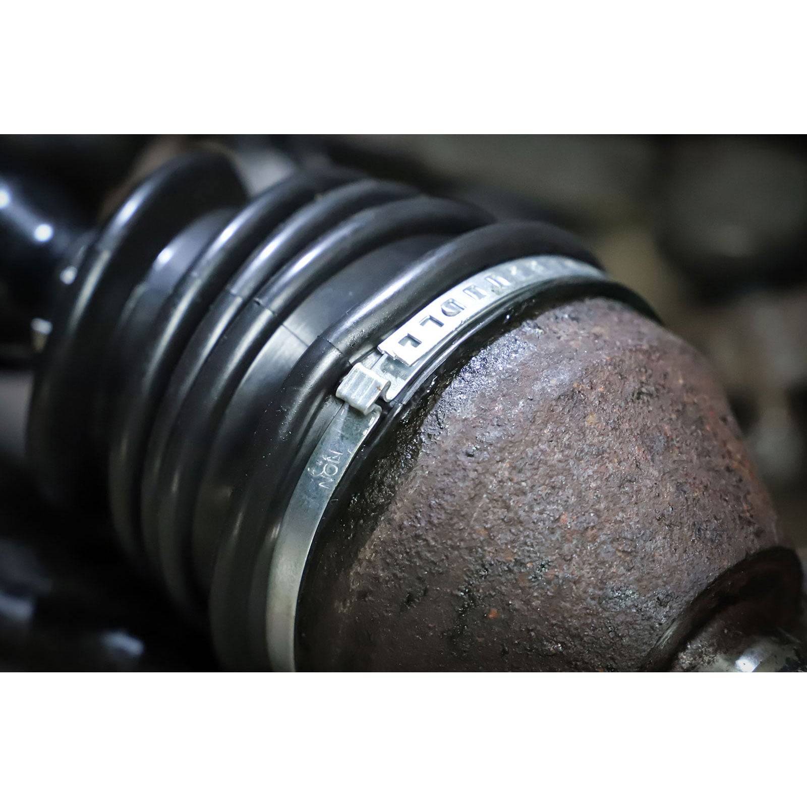 Nahaufnahme eines Autoteils mit einer verrosteten Metalloberfläche und einer schwarzen, akkordeonartigen Gummimanschette, die mit einer Metallschelle befestigt ist.