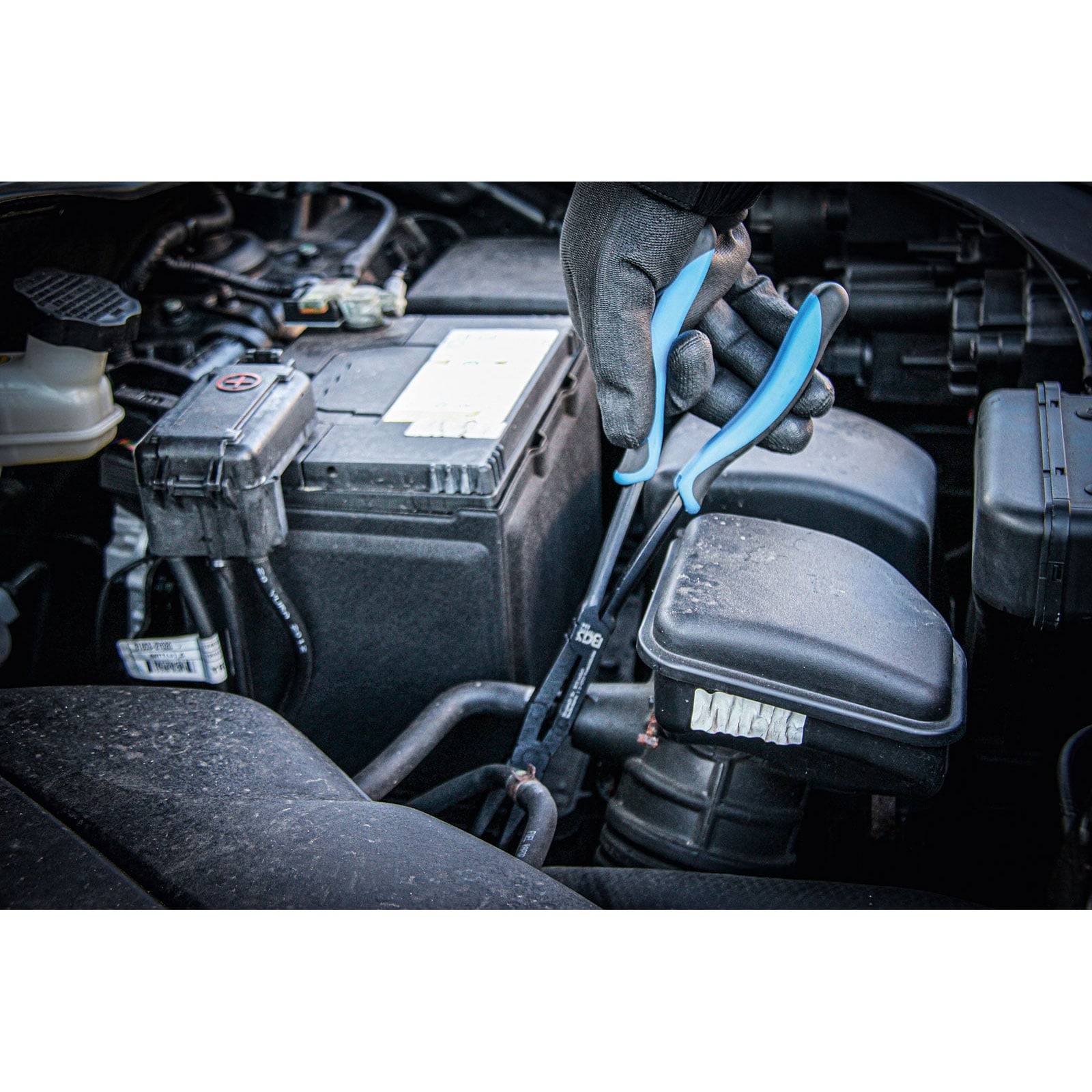 Eine behandschuhte Hand verwendet eine Zange in der Nähe einer Autobatterie im Motorraum und konzentriert sich auf Wartungs- oder Reparaturarbeiten.