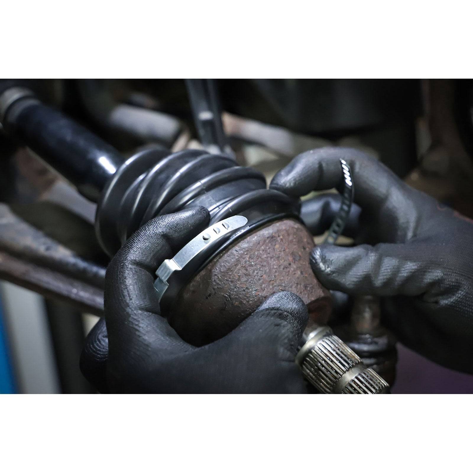 Hände mit schwarzen Handschuhen arbeiten an der Kardanwelle eines Fahrzeugs und justieren eine Metallschelle. Das Bild konzentriert sich auf eine Autoreparatur.