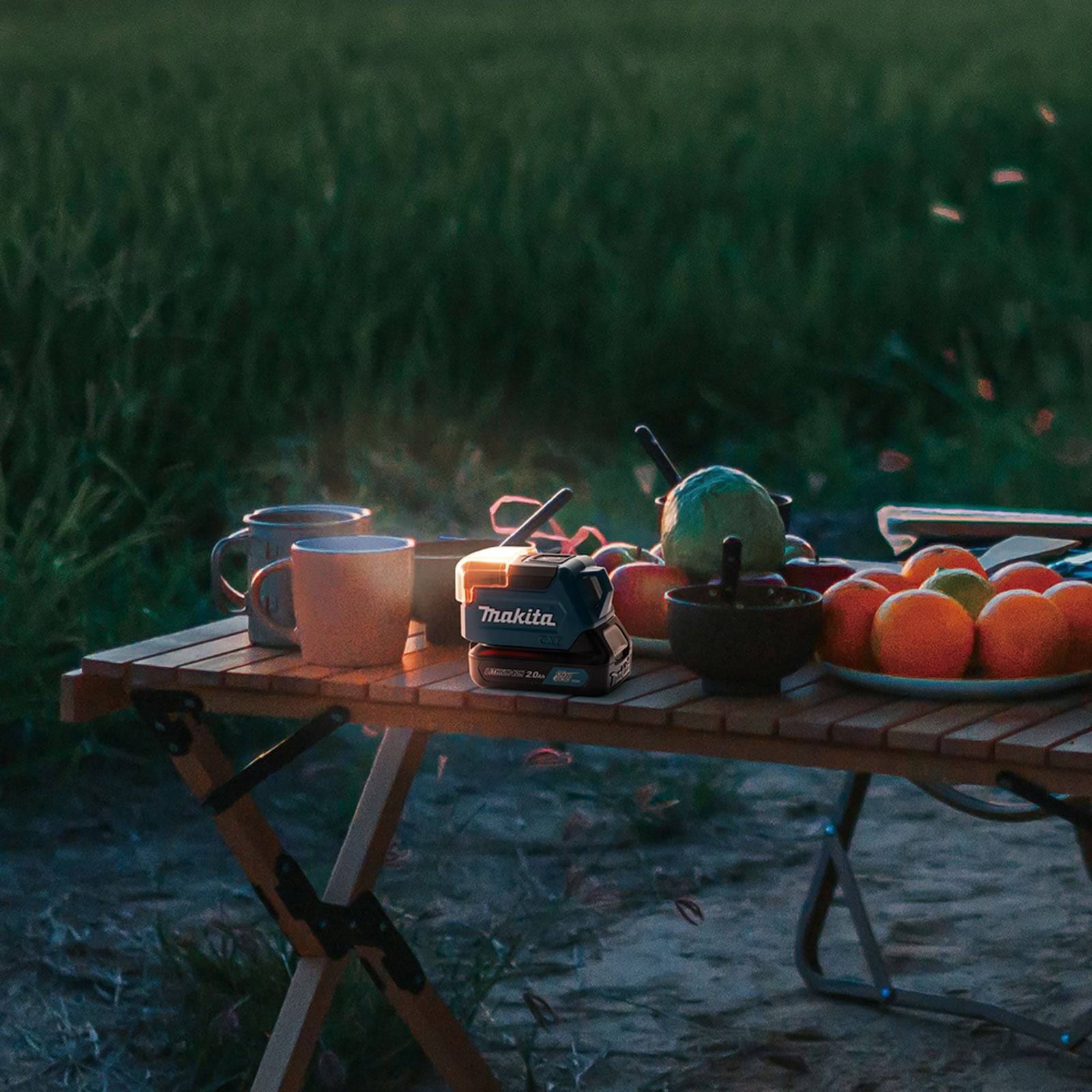 Ein Holztisch mit einer Bohrbatterie, Kaffeetassen, einer Schüssel mit Äpfeln und Obst in einer Feldumgebung im Freien, der eine entspannte Picknick-Atmosphäre vermittelt.