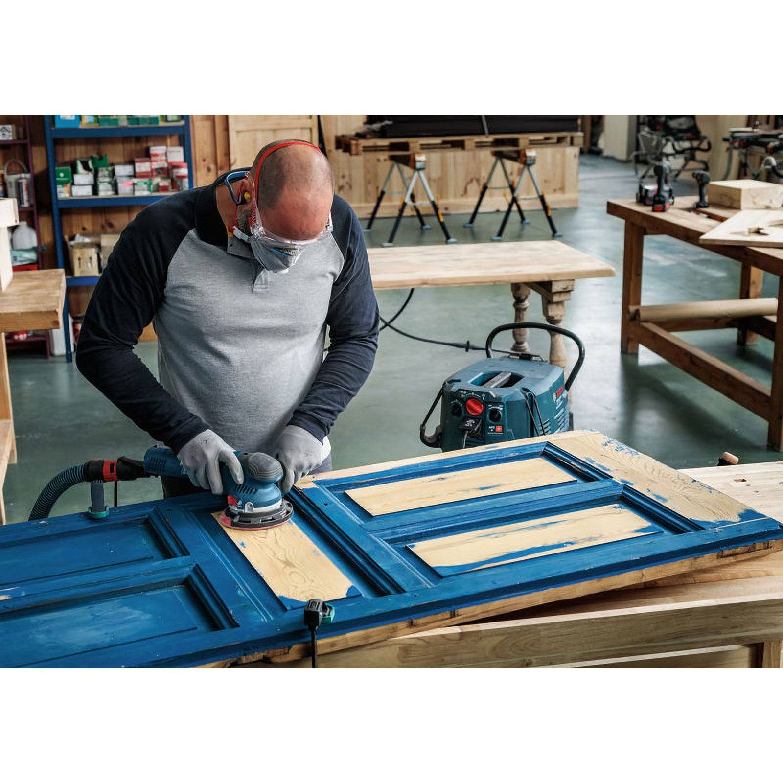 Eine Person mit Sicherheitsausrüstung beim Schleifen einer blauen Holztür, die auf einem Tisch in einer Werkstatt liegt, umgeben von verschiedenen Werkzeugen und Holzgegenständen.