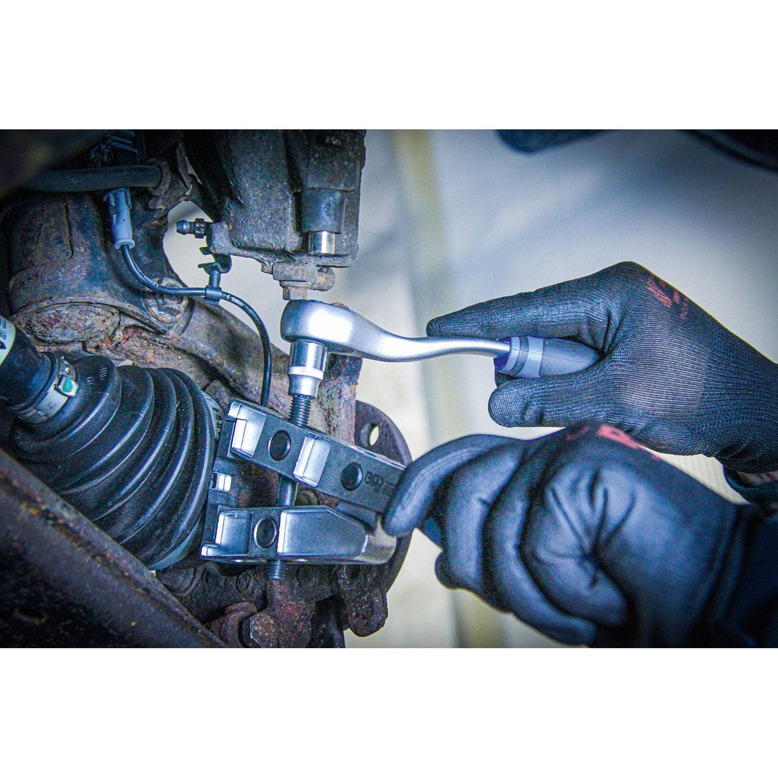 Nahaufnahme von behandschuhten Händen, die ein Werkzeug verwenden, um eine Metallkomponente unter einem Fahrzeug anzupassen, wobei Kraftfahrzeugreparaturarbeiten demonstriert werden.