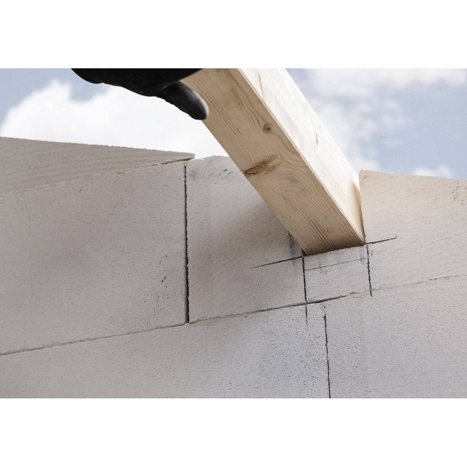 Eine Handschuh tragende Hand hält ein Holzbrett, um Schnitte auf gestapelten Betonblöcken auszurichten und zu markieren, vor einem bewölkten Himmel.