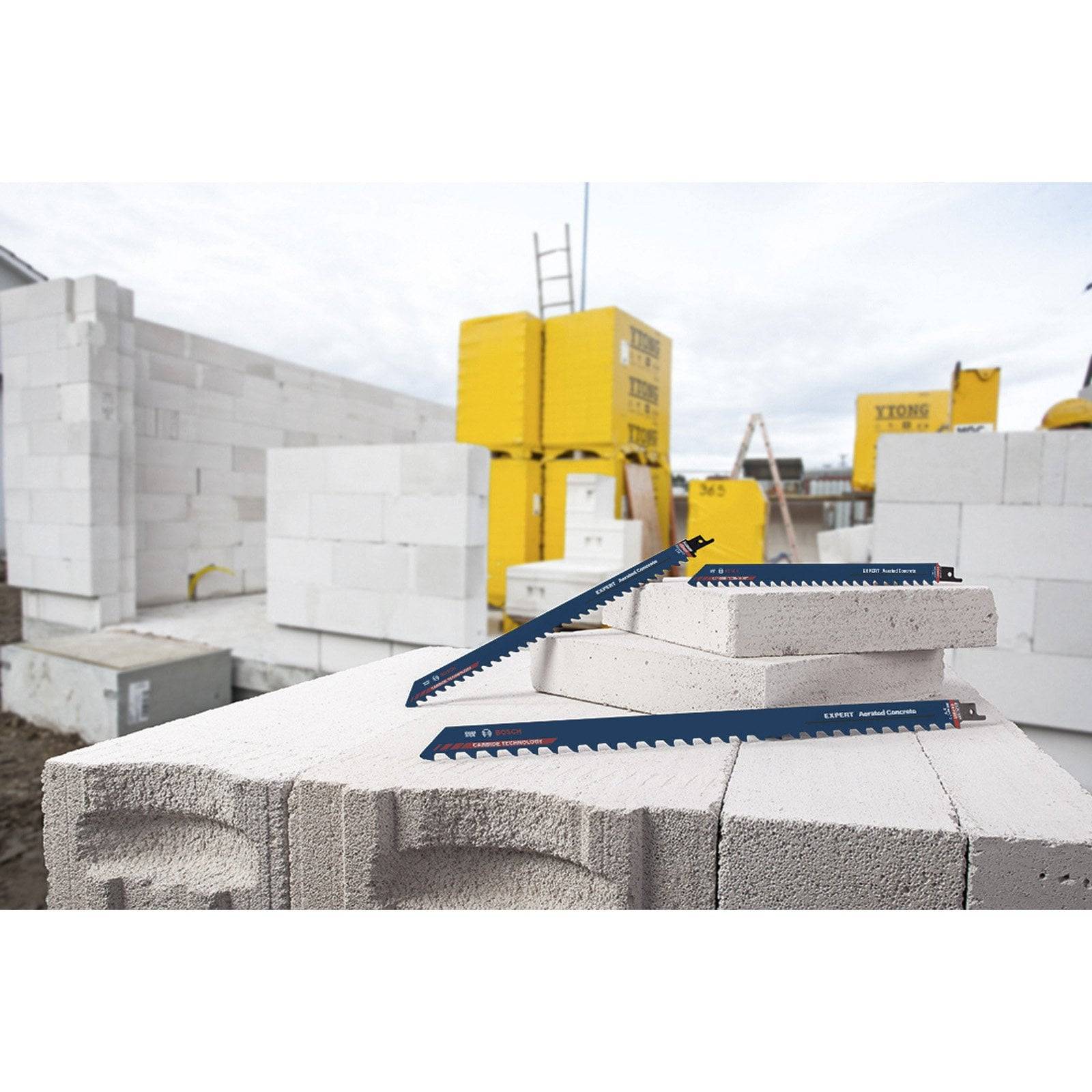 Baustelle mit gestapelten weißen Ziegelsteinen, gelber Baumaschine im Hintergrund und zwei Messwerkzeugen auf einem Ziegelstein im Vordergrund.