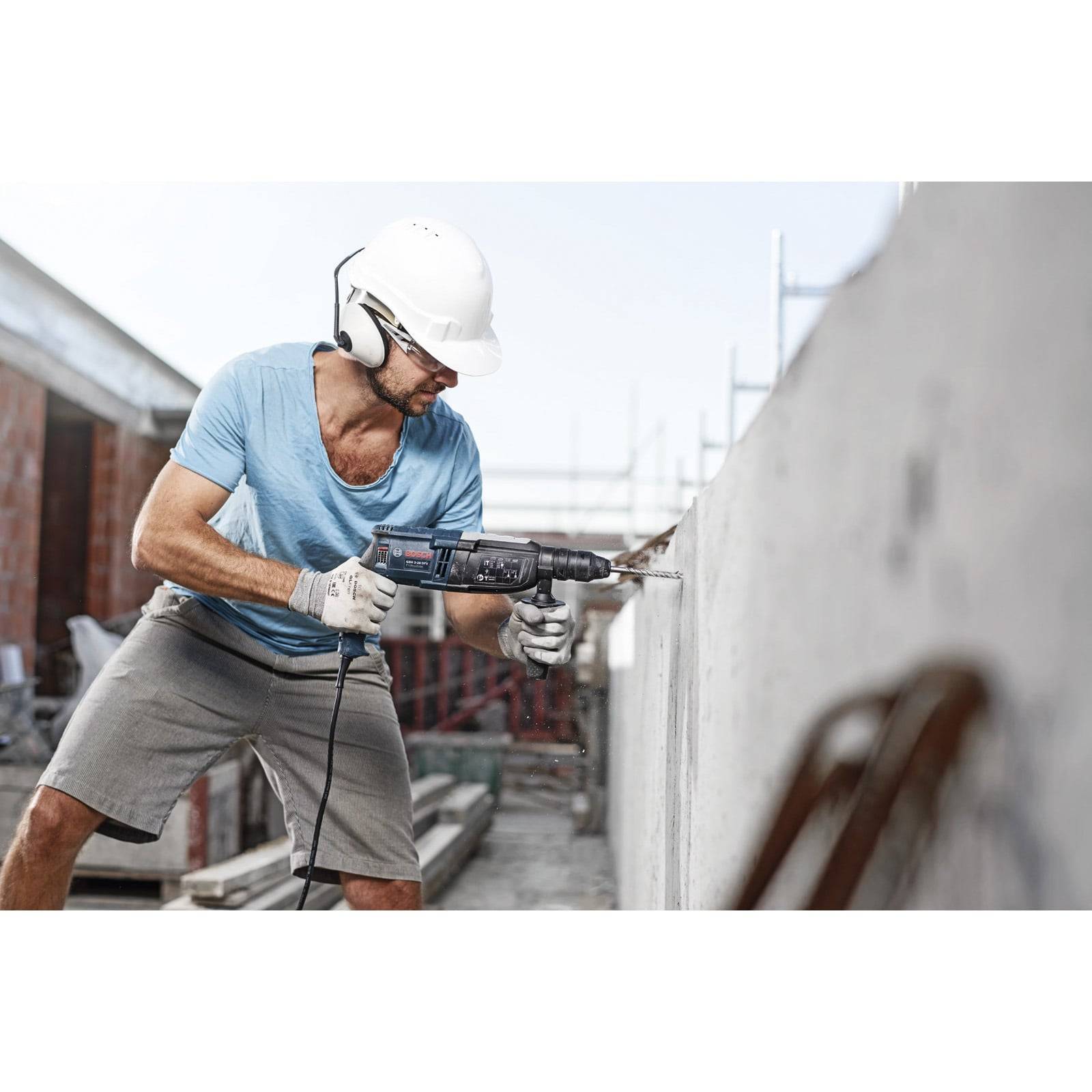 Ein Bauarbeiter bohrt in eine Betonwand an einer Baustelle und trägt dabei einen weißen Schutzhelm und Sicherheitshandschuhe. Gerüste sind zu sehen.