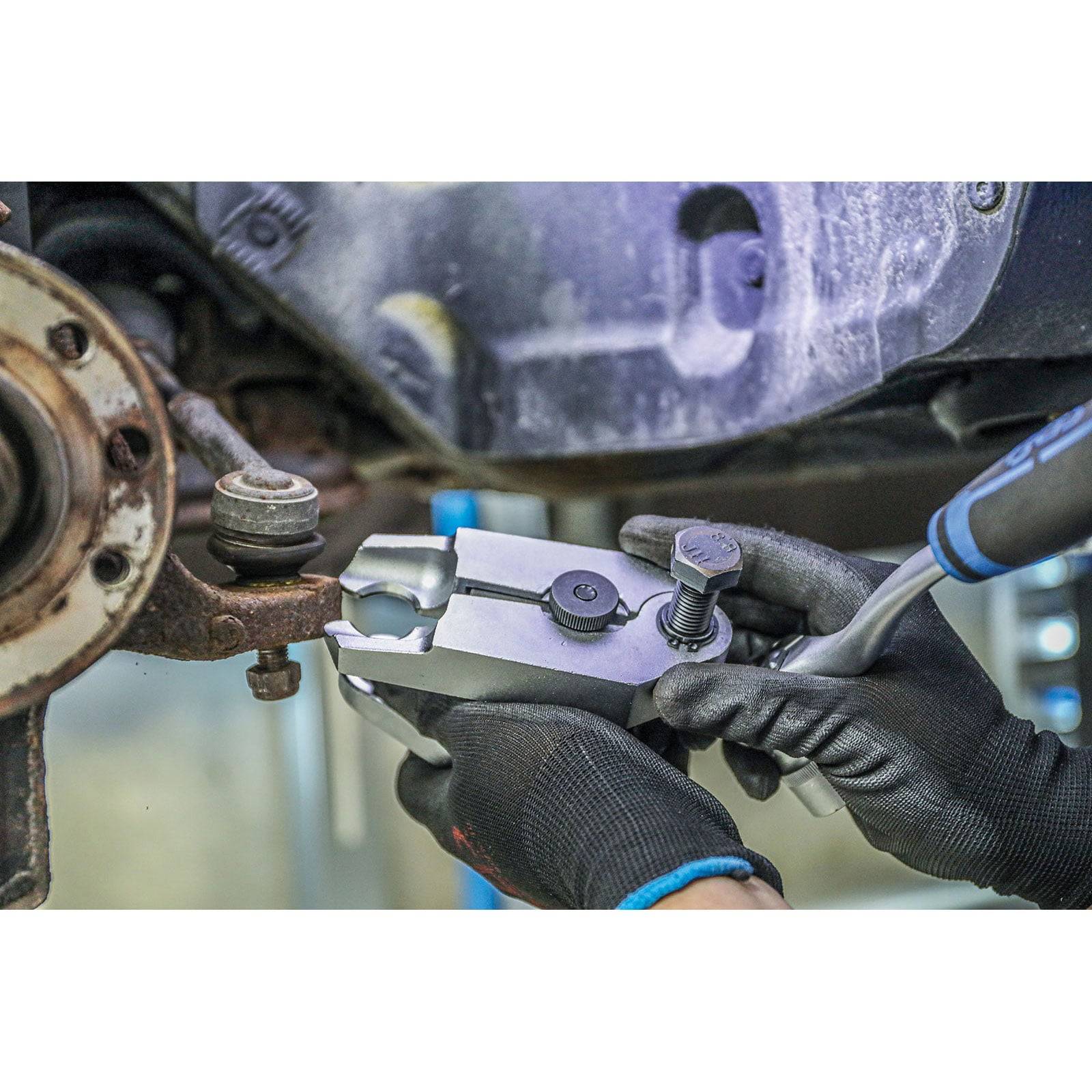 Mechaniker verwendet ein Kugelgelenk-Trennwerkzeug an einer Fahrzeug-Fahrwerkskomponente und trägt dabei schwarze Schutzhandschuhe.