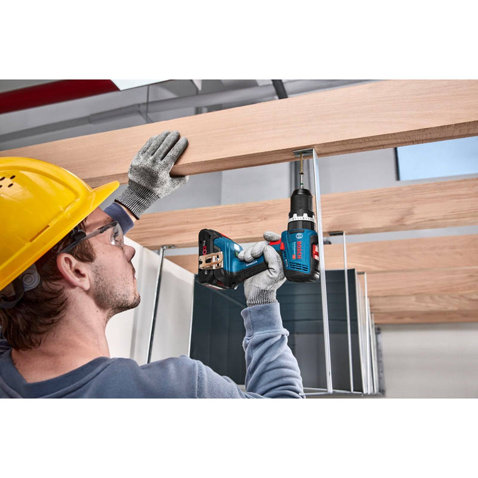 Eine Person mit einem gelben Schutzhelm und Sicherheitsbrille verwendet ein Elektrowerkzeug, um einen Holzbalken an seinem Platz auf einer Baustelle zu befestigen.
