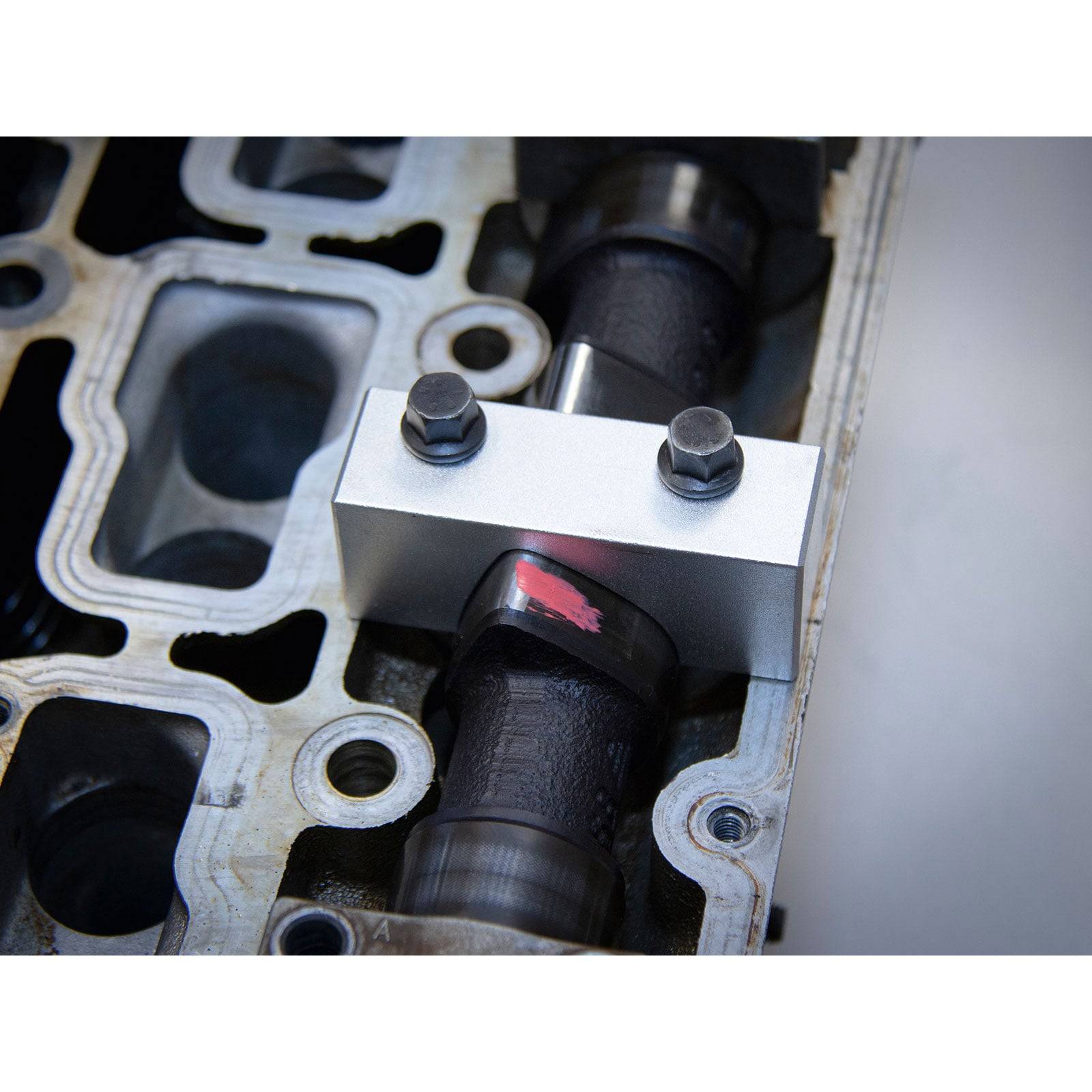Das Bild zeigt eine Metallnockenwelle in einem Motorblock mit zwei Schrauben, die eine Halterung oben befestigen. Ein roter Markierungspunkt ist auf der Nocke der Nockenwelle sichtbar.