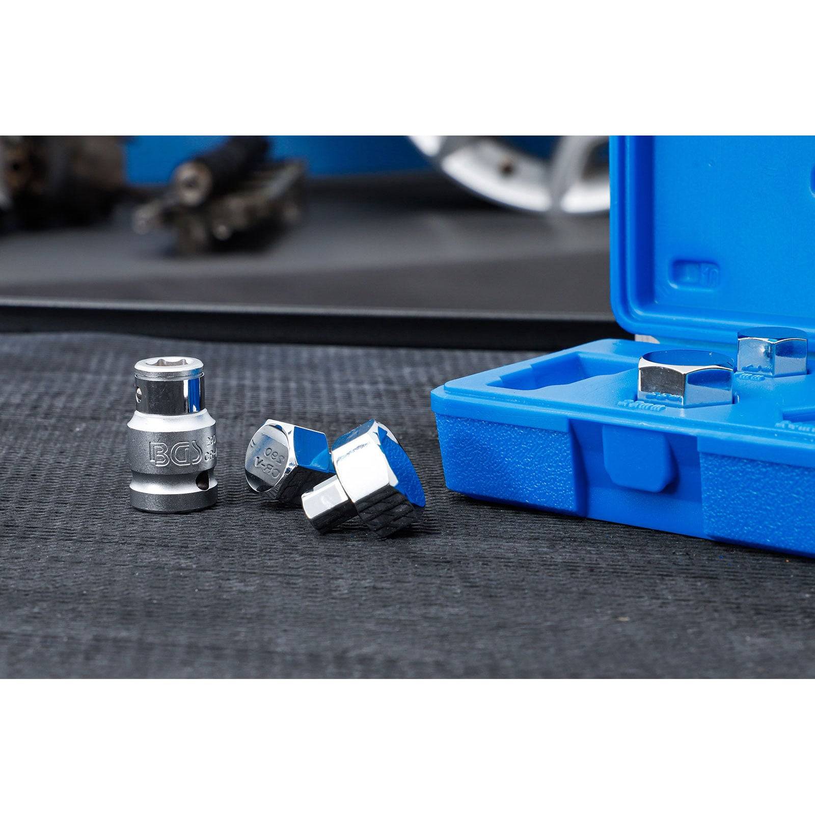 Silberne Metallsockelteile werden neben einer offenen blauen Plastikbox auf einer strukturierten Oberfläche ausgelegt, mit unscharfen Werkzeugen im Hintergrund.