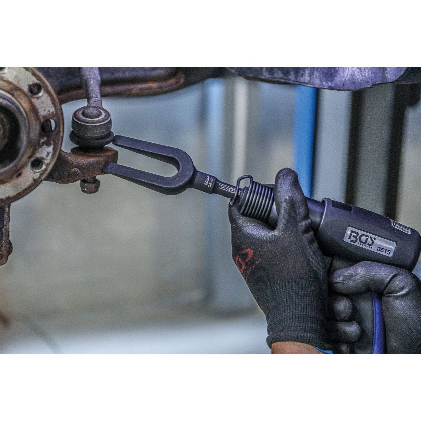 Eine Person verwendet ein Werkzeug, um das Kugelgelenk eines Autos vom Federsystem zu trennen. Handschuhe schützen ihre Hände. Das Werkzeug und das Gelenk sind im Fokus.