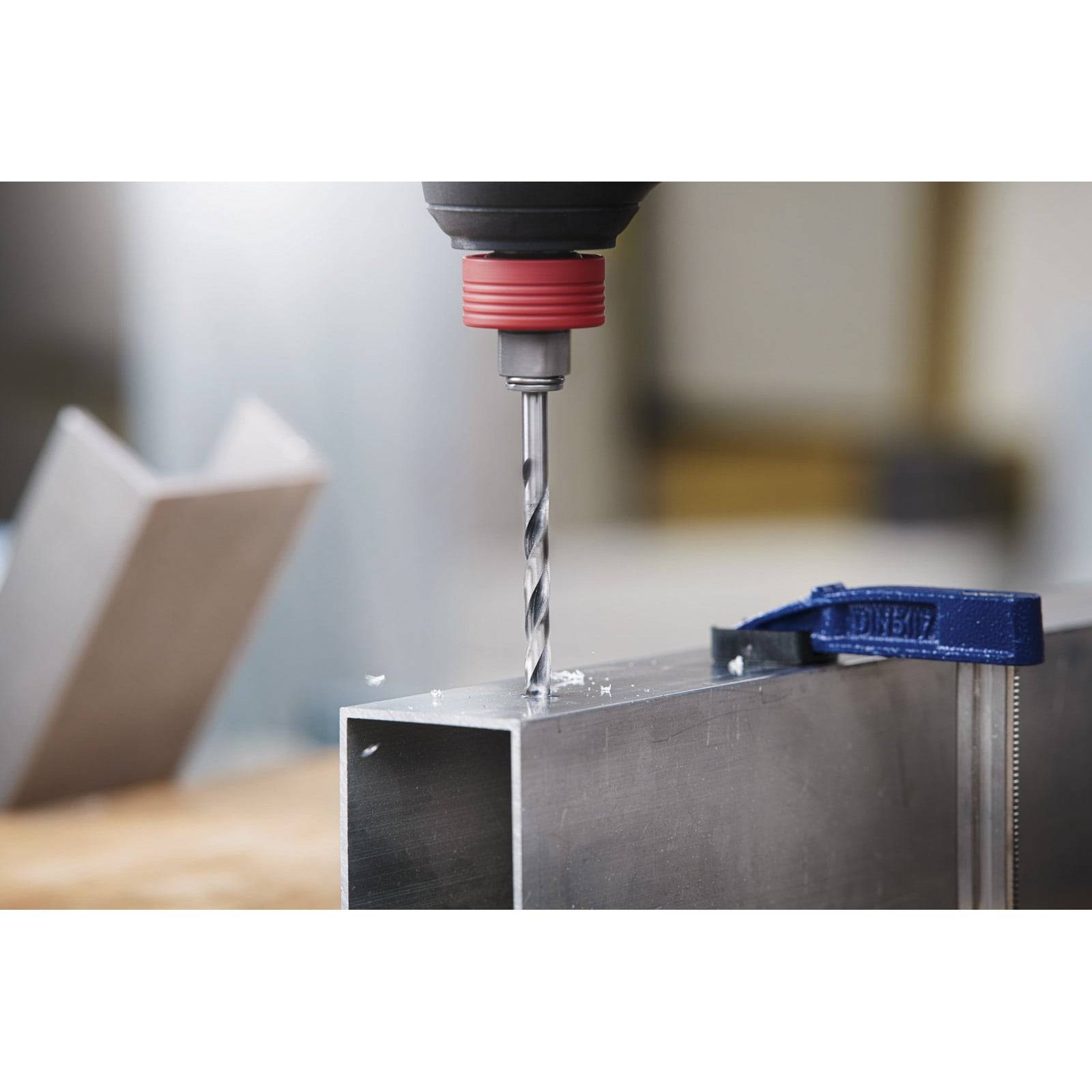 Eine Bohrmaschine, die ein Loch in einen Metallträger bohrt, mit Metallspänen um den Bohrer herum. Ein Metallhalter ist rechts an dem Träger geklemmt.