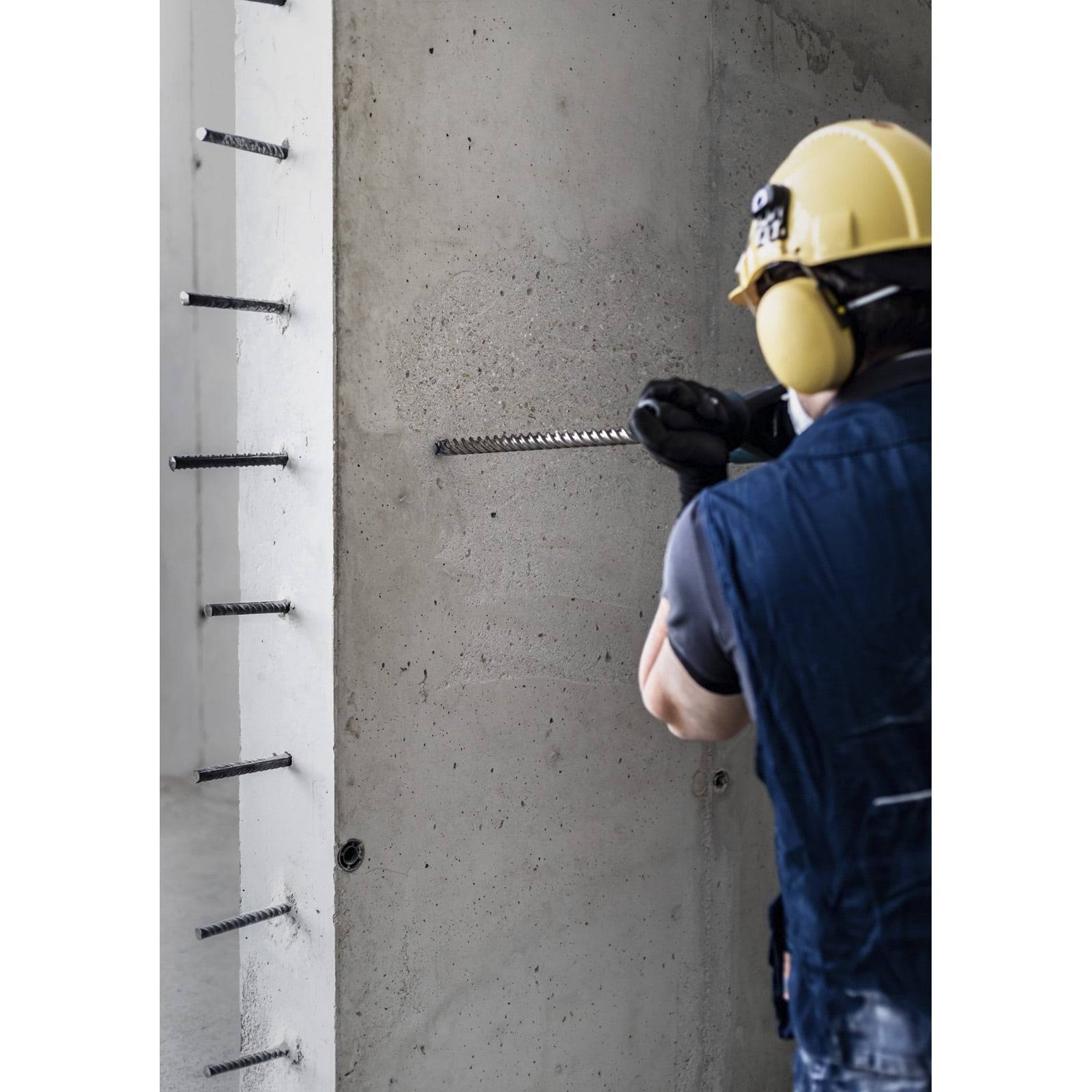 Ein Arbeiter in Schutzausrüstung schiebt eine Stahlstange in eine Betonwand, umgeben von anderen eingelassenen Stäben, was auf Bau- oder Verstärkungsarbeiten hindeutet.