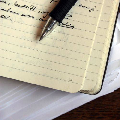 Nahaufnahme eines Stifts, der auf einem offenen Notizbuch mit Linien ruht, auf dem handschriftliche Notizen sichtbar sind, mit einer Seitenkante und Schreibtischoberfläche.