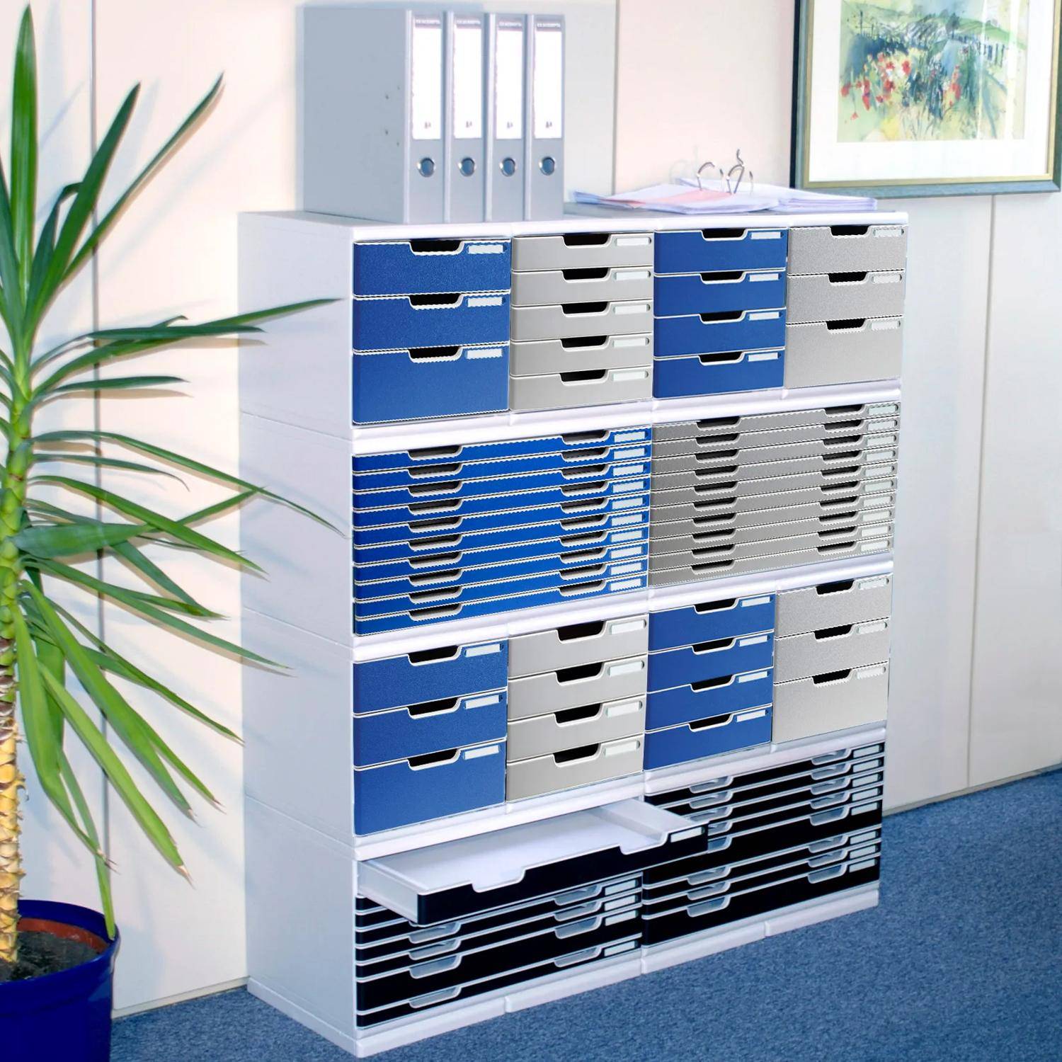 Hohes weißes Büro-Lagersystem mit vielen blauen, grauen und schwarzen Dokumentablageboxen und Ordnern, neben einer Topfpflanze und Wandkunst.
