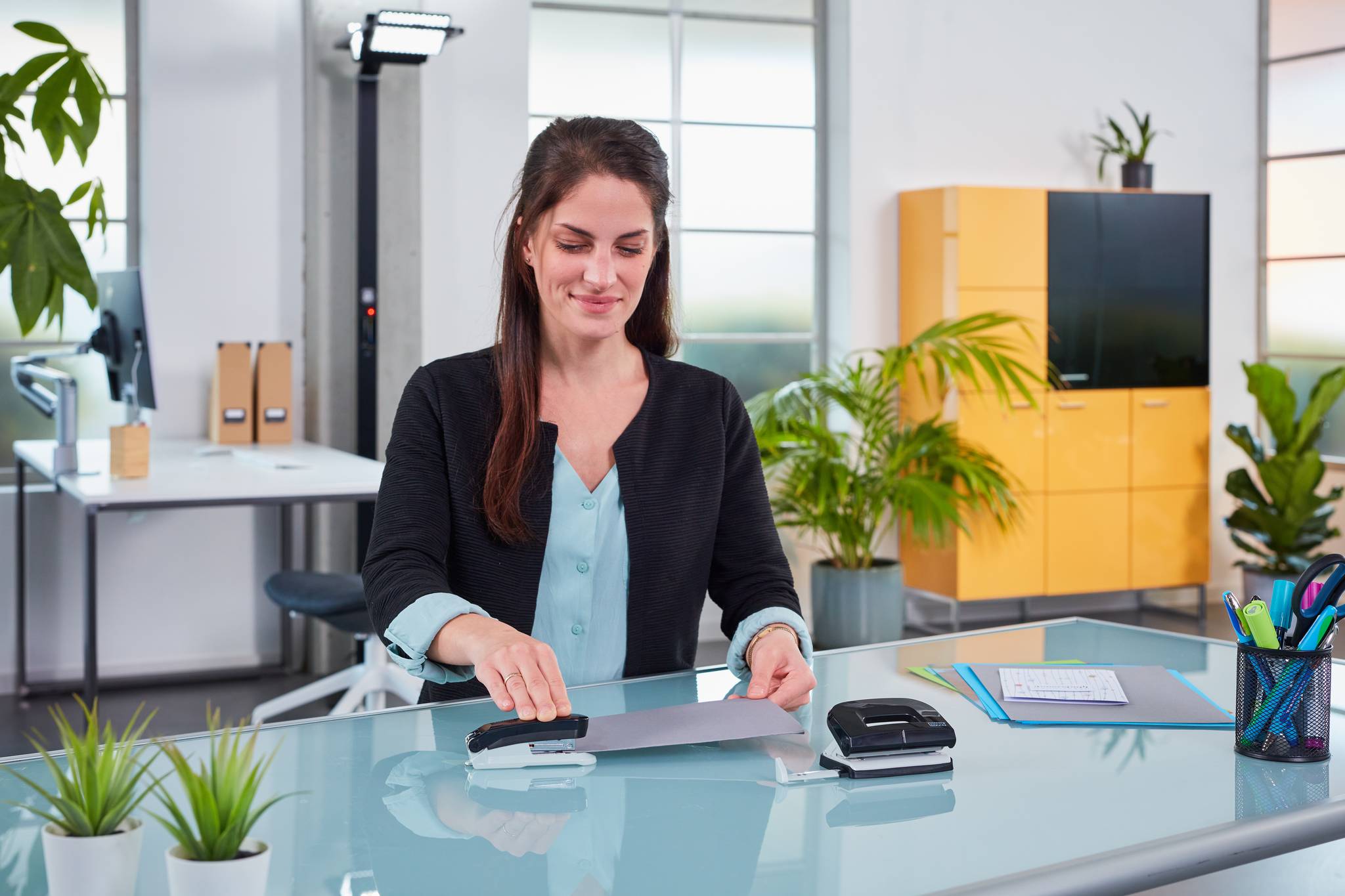 Eine Frau in einem Büro laminiert ein Blatt an einem Glastisch, mit Büropflanzen, Schränken und Materialien im Hintergrund.