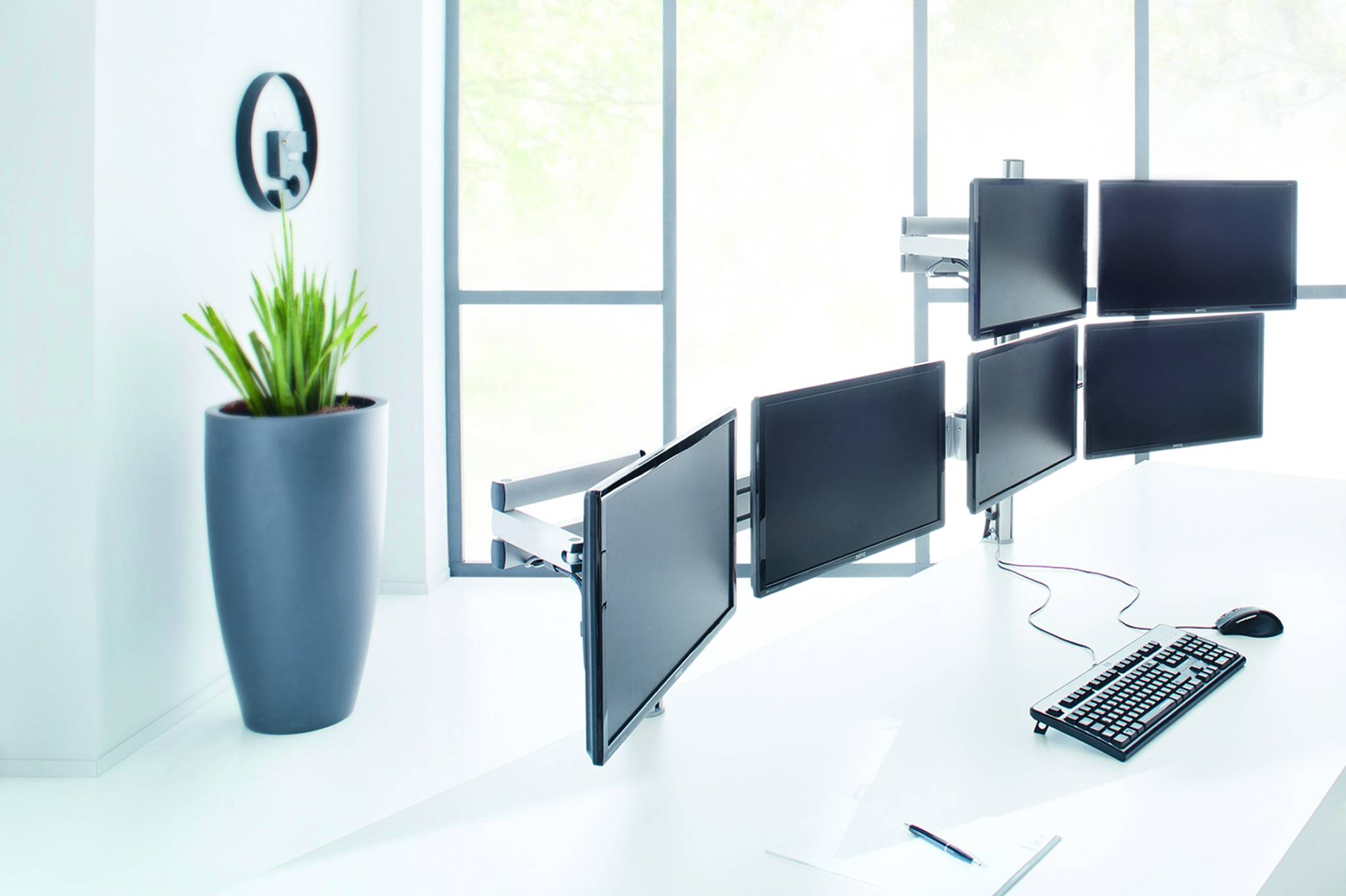 Moderner weißer Bürotisch mit mehreren angebrachten Computermonitoren, Tastatur, Maus und einer Topfpflanze auf der linken Seite.