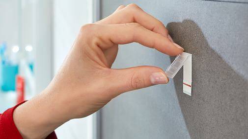 Eine Hand, die einen kleinen Klebestreifen oder eine Registerkarte auf einer grauen Wand oder Gerätoberfläche anbringt.