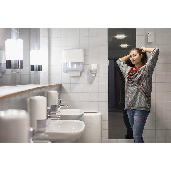 Frau, die Kopfhörer anzieht, während sie in einem gefliesten öffentlichen Badezimmer neben Waschbecken und Spiegeln steht