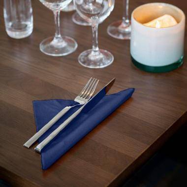 Blaue Serviette, um eine Gabel und ein Messer auf einem Holztisch gefaltet, mit Weingläsern und einer brennenden Kerze in der Nähe.