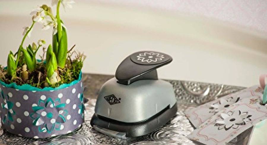 Handgestanztes Handwerkzeug mit blumenförmigem Ausschnitt neben einem dekorierten Blumentopf und passenden gestanzten Papiermustern auf einem Tisch.