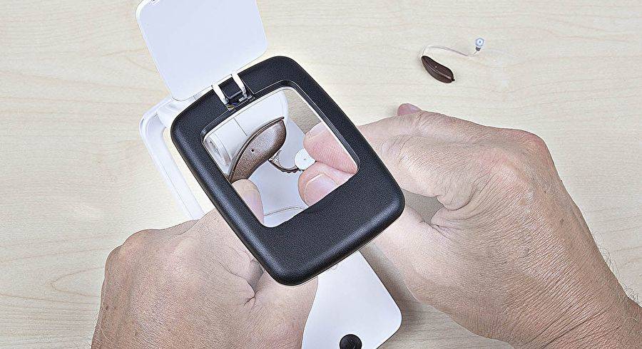 Hände halten ein Hörgerät, während eine kleine Batterie in sein offenes Fach auf einem Tisch eingesetzt wird.