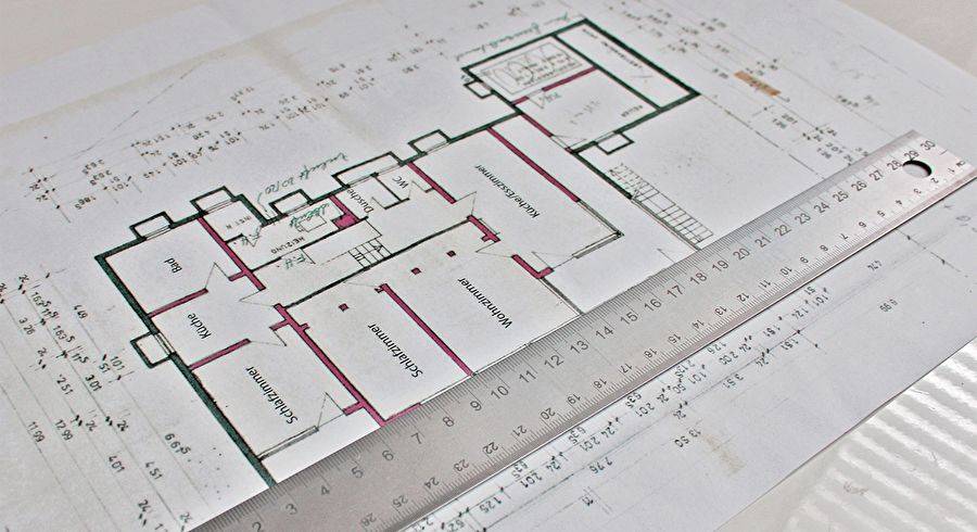 Architektonischer Grundriss auf Papier mit Raumbezeichnungen und Maßstäben, überlagert durch ein Stahllineal als Maßstab.