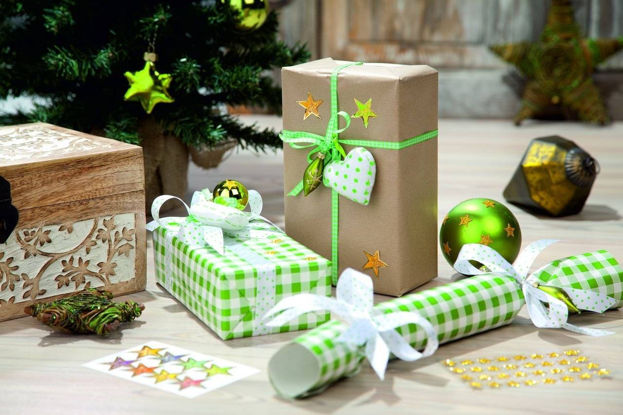 Eingepackte Weihnachtsgeschenke und Dekorationen auf einem Tisch neben einem beleuchteten Baum, einschließlich Schachteln, Schleifen, Stern-Ornamente und einer Geschenktüte.