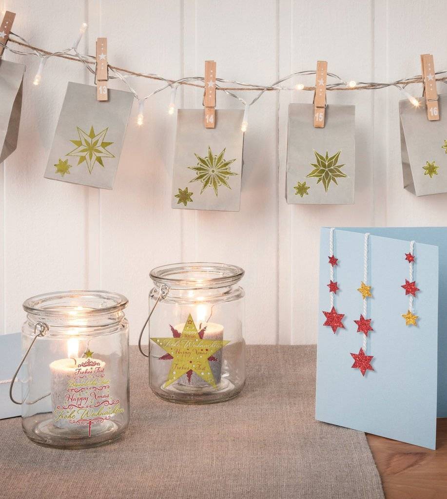 Handgefertigte Weihnachtskarten und Geschenkgläser mit brennenden Kerzen, verziert mit sternförmigen Ornamenten und Lichterketten auf einem Tisch.