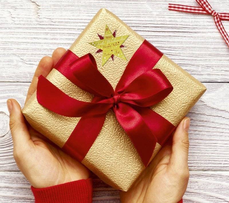 Hände halten eine in Gold eingewickelte Geschenkbox mit einer roten Schleife und einem Sternetikett auf einer hellen Holzoberfläche.